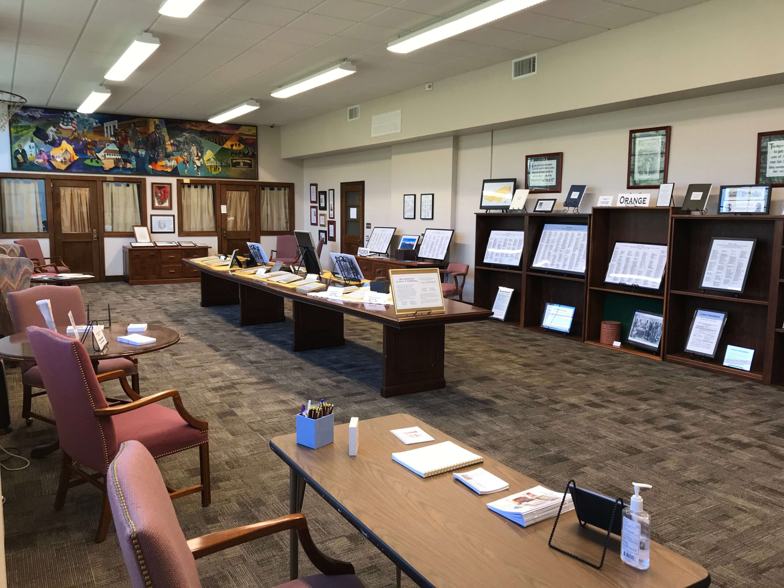 A spacious room features a large wooden table at the center displaying various items and documents. Surrounding shelves are filled with framed certificates and documents. There are chairs and smaller tables with office supplies, and a mural adorns one wall.