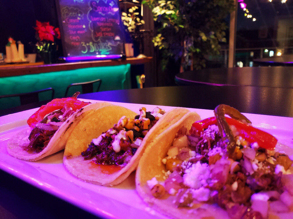 A close-up of three tacos on a white plate, each filled with different toppings like grilled vegetables, cheese, and meat. In the background, there's a neon-lit bar area with a chalkboard menu, vibrant decor, and string lights creating a cozy ambiance.