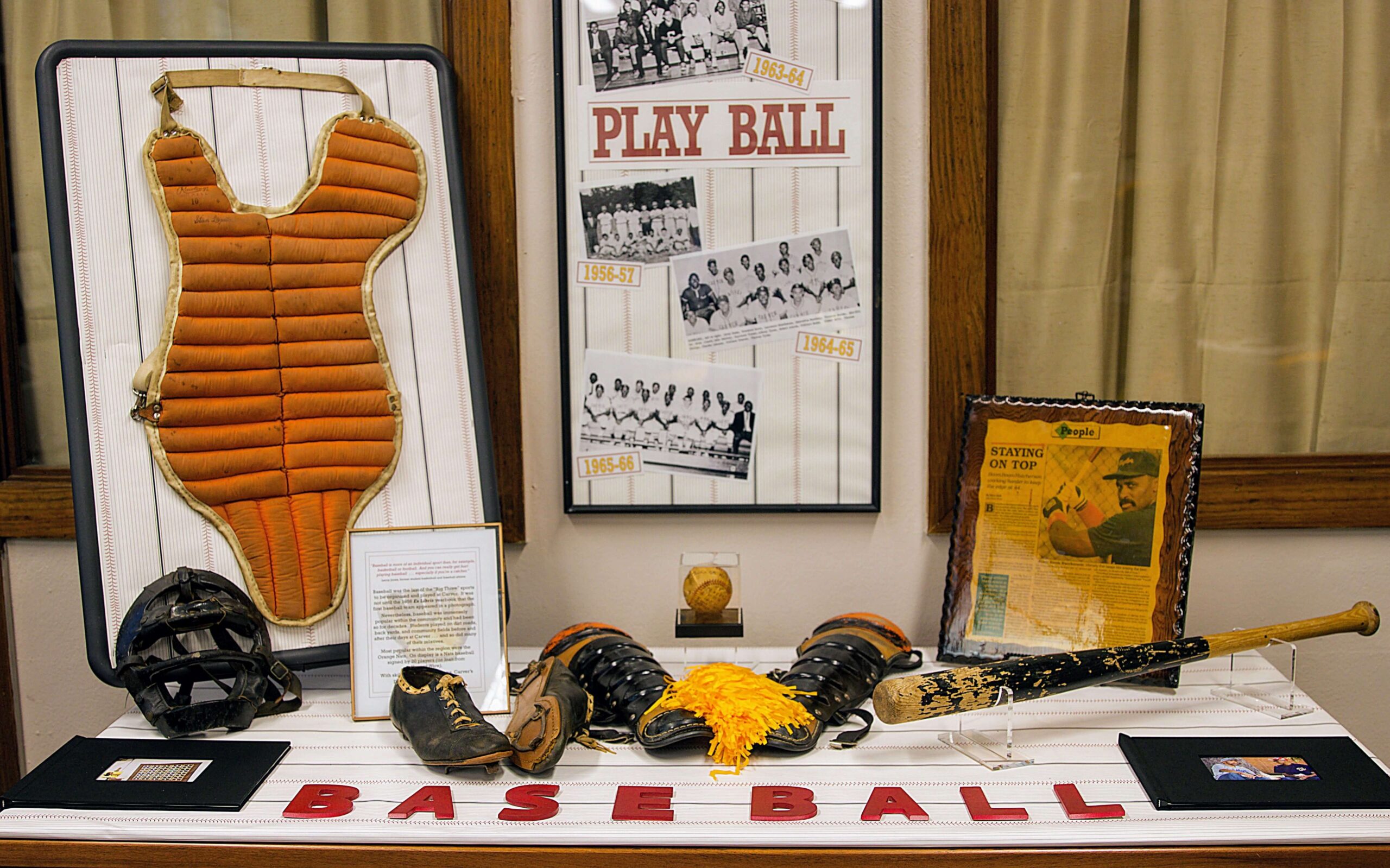 A museum display featuring vintage baseball memorabilia. Items include a chest protector, catcher's mask, gloves, baseballs, a bat, and cleats. Behind the display, there are framed photos, including a 