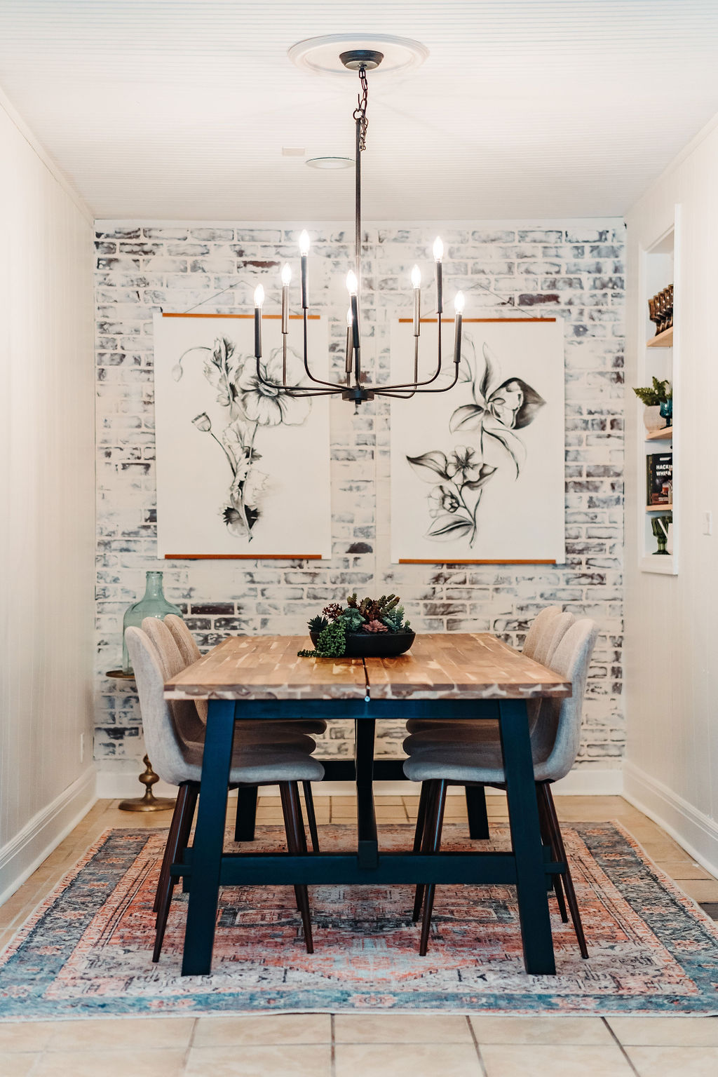 A cozy dining room with a wooden table and four upholstered chairs, featuring a modern chandelier. The wall behind the table displays two abstract line art pieces on a white brick wall. A colorful rug enhances the warm atmosphere.