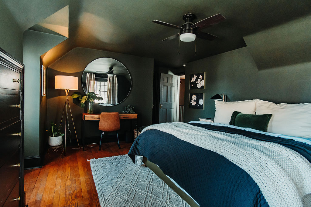 A cozy bedroom features dark green walls, a large bed with white and navy bedding, and a round mirror above a wooden desk. A brown chair sits at the desk, and a standing lamp and potted plant add warmth to the room. Two framed pictures hang above the bed.