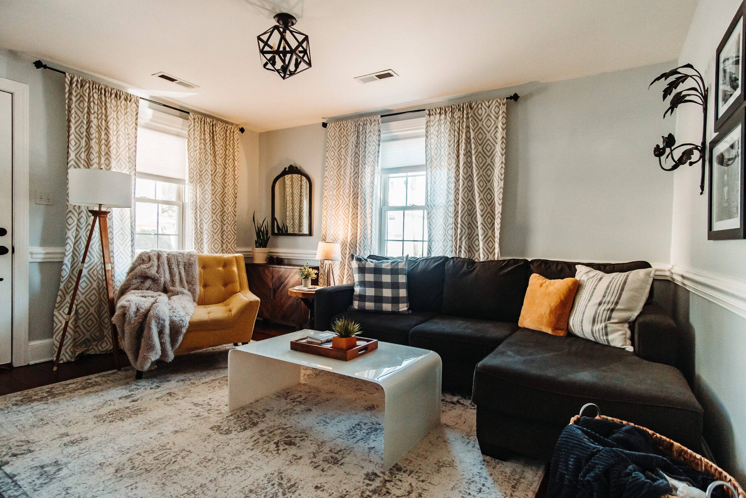 A cozy living room with a sectional sofa adorned with pillows and a blanket, a yellow armchair with a throw, and a modern white coffee table. The room features two windows with patterned curtains, a floor lamp, wall art, and a potted plant, creating a welcoming atmosphere.