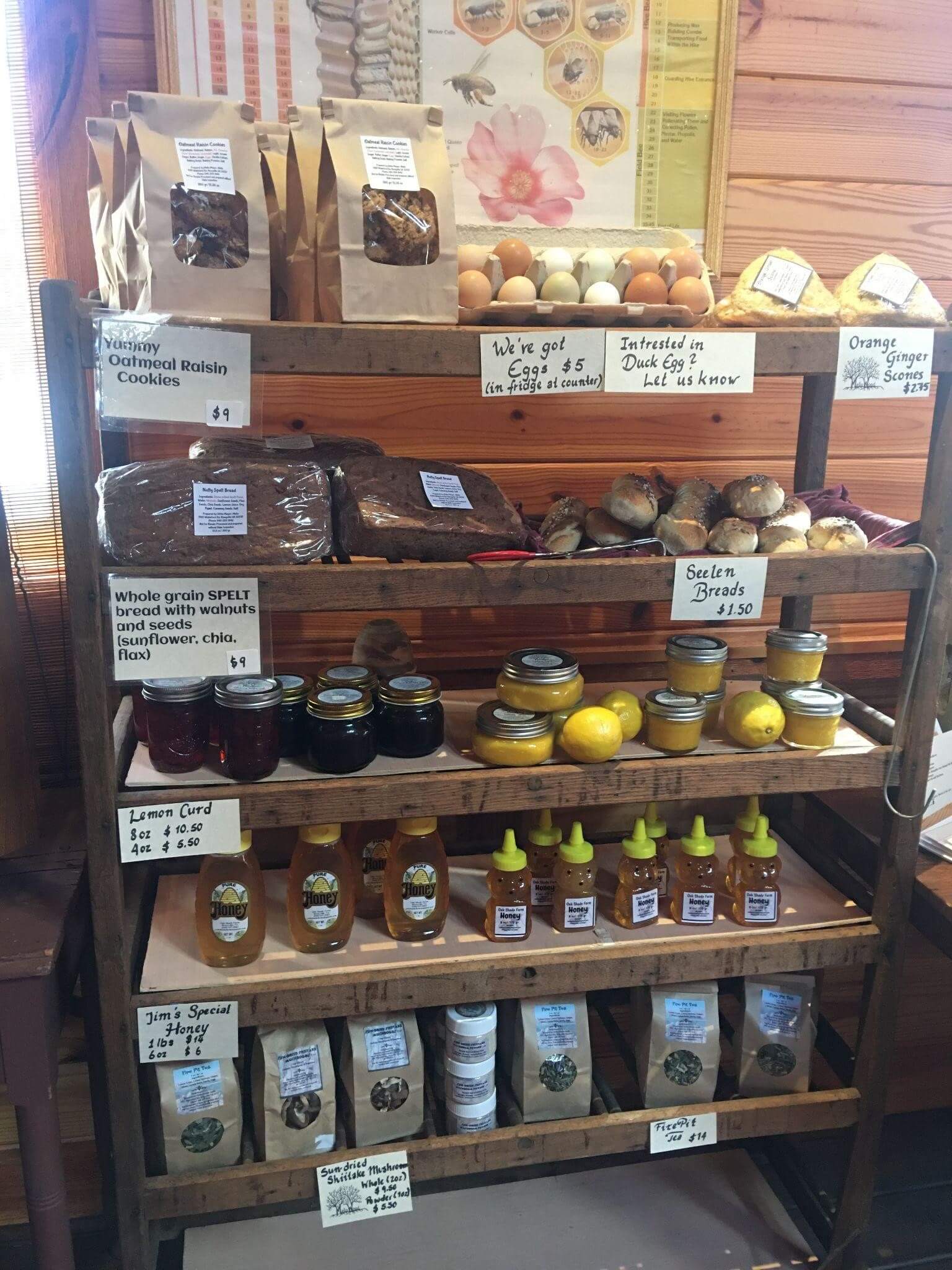 A wooden shelf in a farm store displaying assorted goods. Top shelves have baked goods, cookies, and fresh eggs. Middle shelves feature honey jars and preserves. Bottom shelf holds honey bottles. Handwritten labels with prices and descriptions are visible.