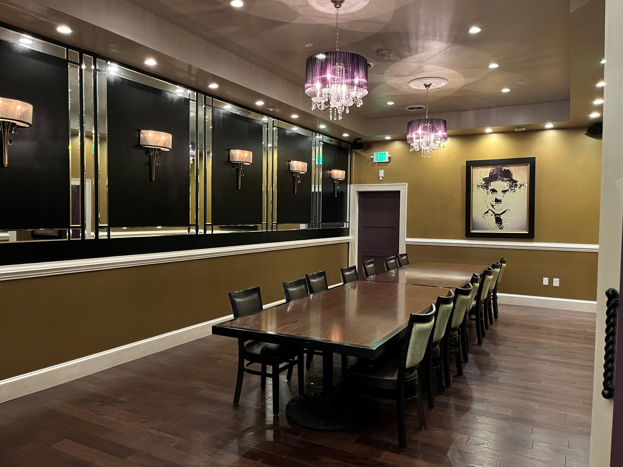 A long, elegant conference room with a polished wooden table surrounded by black chairs. The walls are decorated with black panels, sconces, and a large framed portrait. Three purple chandeliers hang from the ceiling, and there is an exit door at the far end.