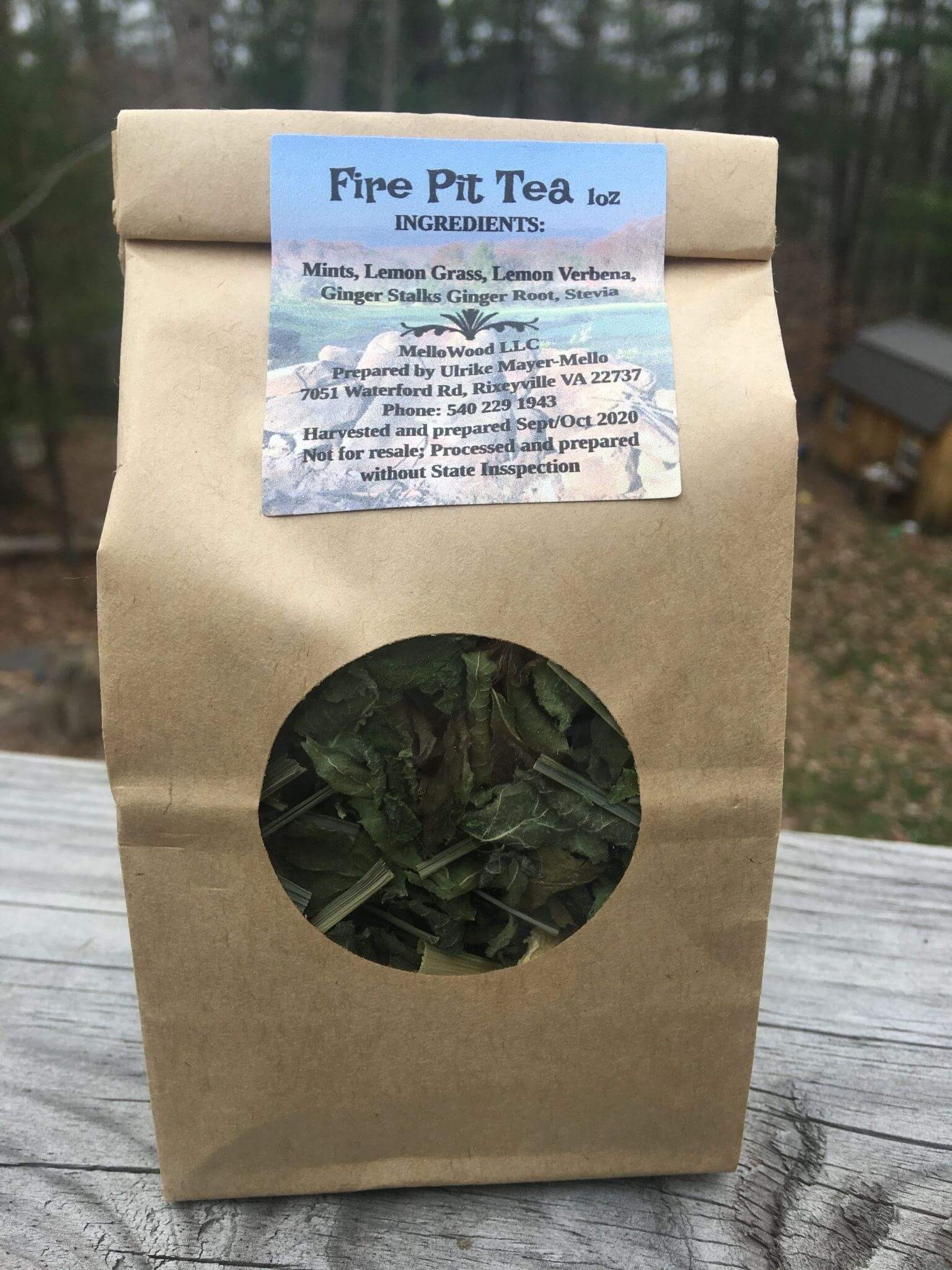 A brown paper package labeled "Fire Pit Tea" contains dried herbs. The label details ingredients: mints, lemon grass, lemon verbena, ginger stalks, ginger root, and stevia. The package is positioned outdoors, with a blurred background of trees and a wooden structure.