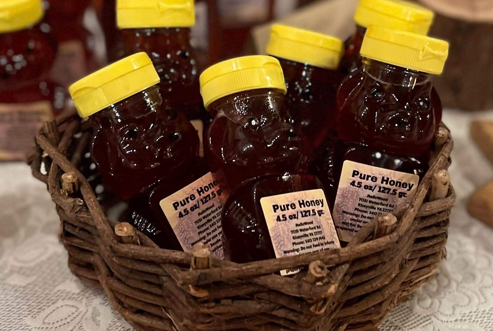 A wicker basket containing several bear-shaped plastic bottles filled with honey. The bottles have yellow lids and labels reading "Pure Honey 4.5 oz / 127.5 gr." The basket is on a lace tablecloth with wooden surfaces and blurry objects in the background.