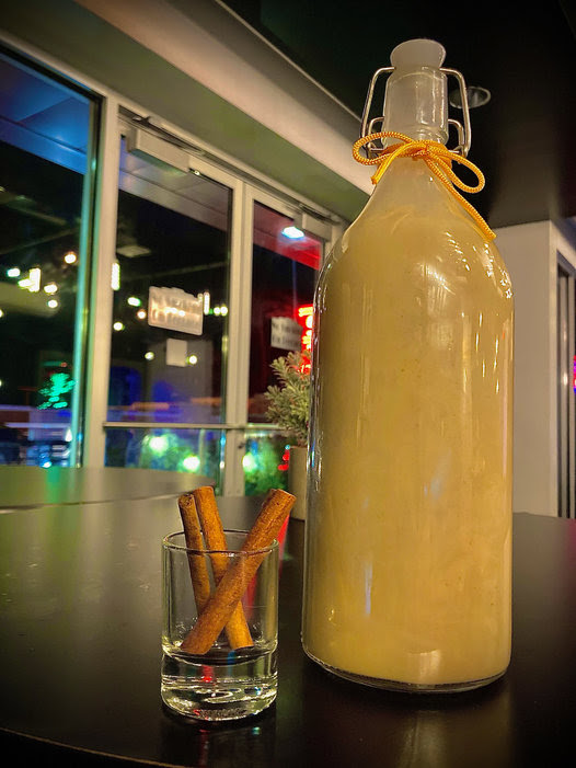 A large glass bottle filled with a creamy yellow beverage is placed on a dark table. Next to it is a small glass containing two cinnamon sticks. The background shows glass windows with colorful lights and reflections.