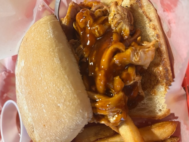 A close-up of a barbecue pulled chicken sandwich served on a bun with crispy golden fries on the side. The sandwich is overflowing with saucy, tender shredded chicken. There are small containers of ketchup and additional sauce in the background.