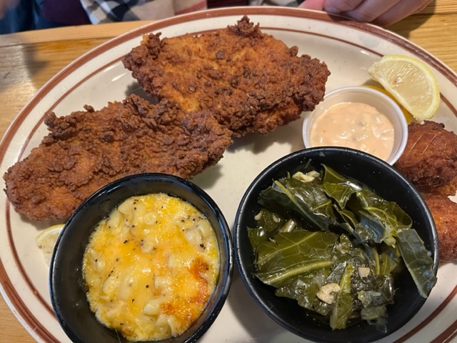 A plate of fried chicken with sides of macaroni and cheese, collard greens, and a small container of dipping sauce. A lemon wedge is also placed on the plate.