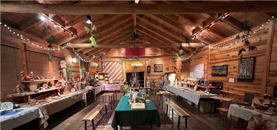 A cozy wooden cabin interior is decorated with string lights and festive ornaments. The room features multiple tables covered with various handcrafted items and decorations. Benches are placed around some of the tables, and the walls are adorned with paintings and crafts.