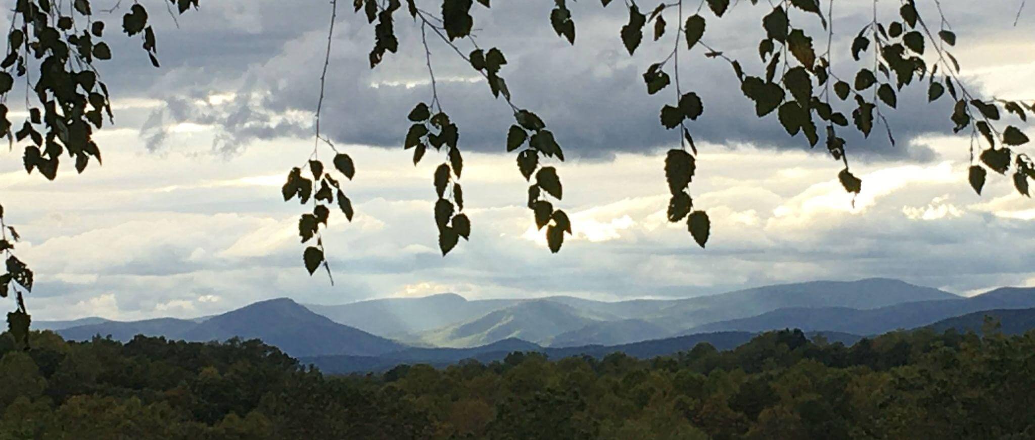 A scenic view of rolling mountains under a cloudy sky. Sun rays peek through gaps in the clouds, casting light on parts of the mountains. Tree branches with leaves frame the top of the image. The foreground has dense, green foliage.