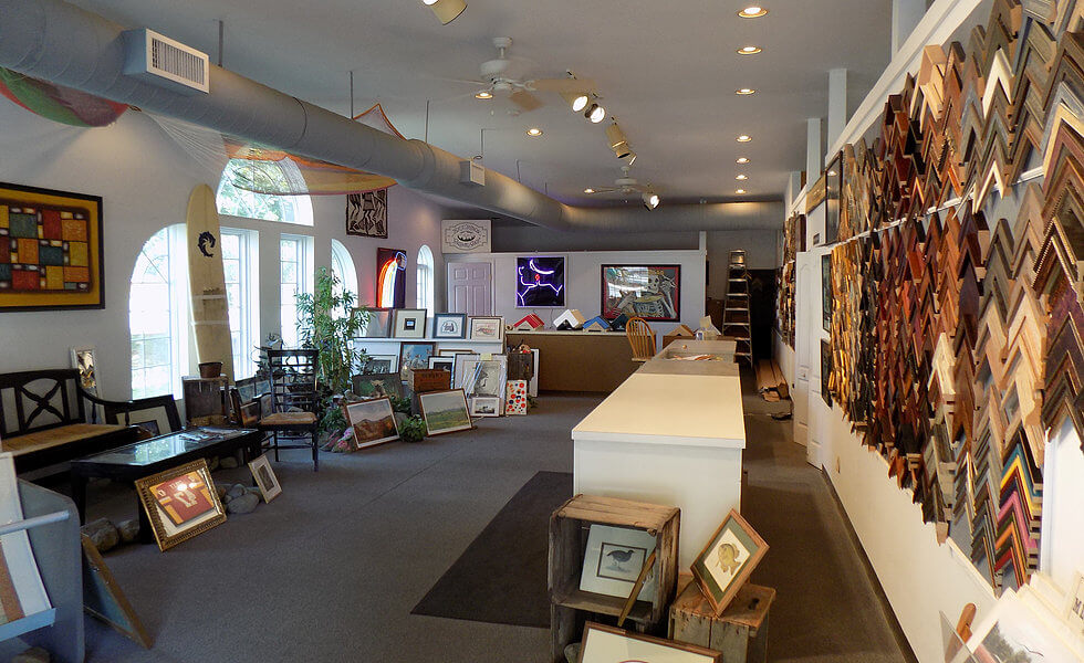 A well-lit art gallery with framed pictures and artwork displayed on the walls and floor. The room features a high ceiling with exposed ductwork and large arched windows on one side. Various photo frames are hung on the walls and placed on trolleys and tables.