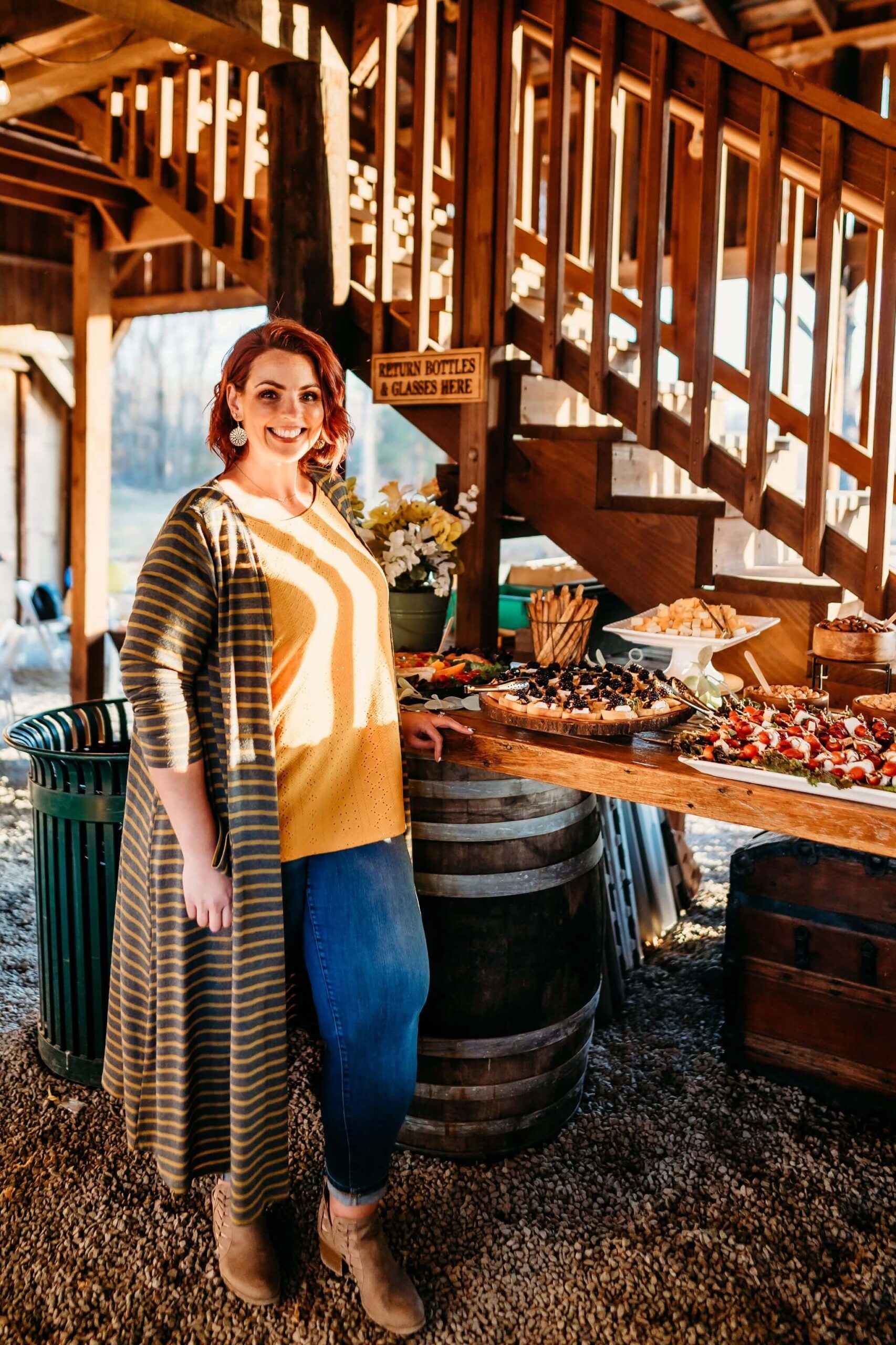 A person with red hair and a smile stands next to a rustic wooden table filled with various plates of food. They are wearing jeans, a yellow shirt, and a long striped cardigan. The setting appears to be a festive outdoor event under a wooden structure.