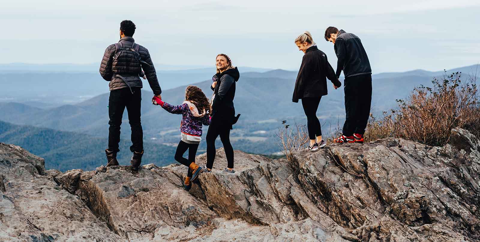 A group of five people, including two adults and three children, stand on a rocky outcrop overlooking a vast mountain range. They are dressed in warm clothing. One child holds hands with an adult while the others look out at the scenic view in the distance.