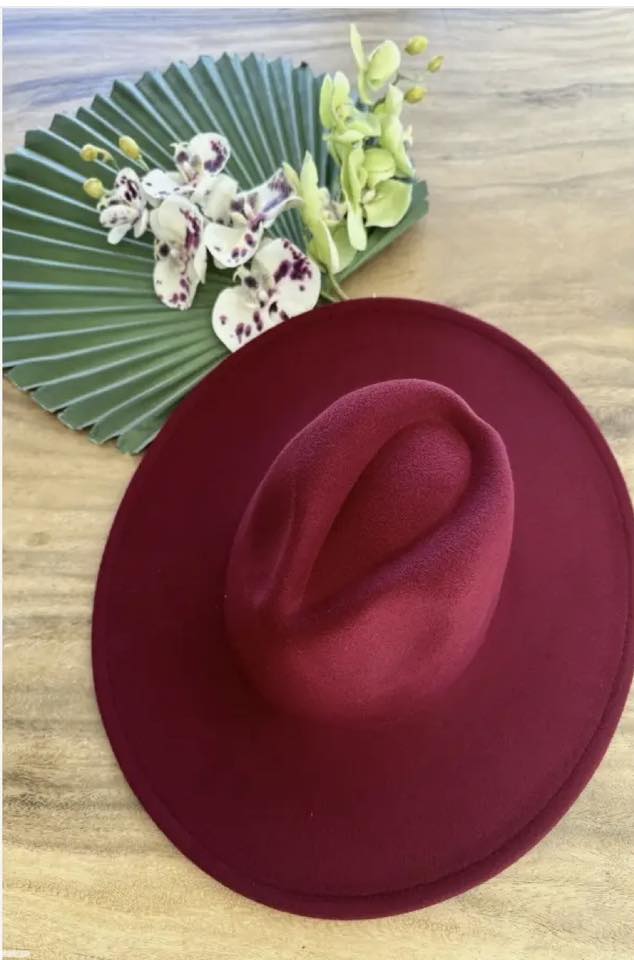 A maroon felt hat lies on a wooden surface beside a green pleated fan and white orchids with purple spots. The arrangement appears elegant and carefully placed, highlighting the contrasting colors and textures.