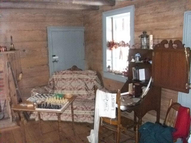 A rustic room with wooden walls and a log ceiling features a vintage patterned sofa, a small table with a chessboard, and an antique wooden desk with a chair. A window with ornate curtains lets in light, highlighting the cozy, old-fashioned ambiance of the space.