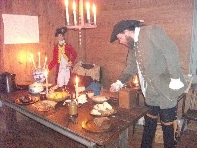 A man dressed in historical 18th-century attire stands by a wooden table with candles and various foods. A cardboard cutout of a colonial soldier decorates the wooden wall behind him. The scene appears to be a reenactment or a historical display.