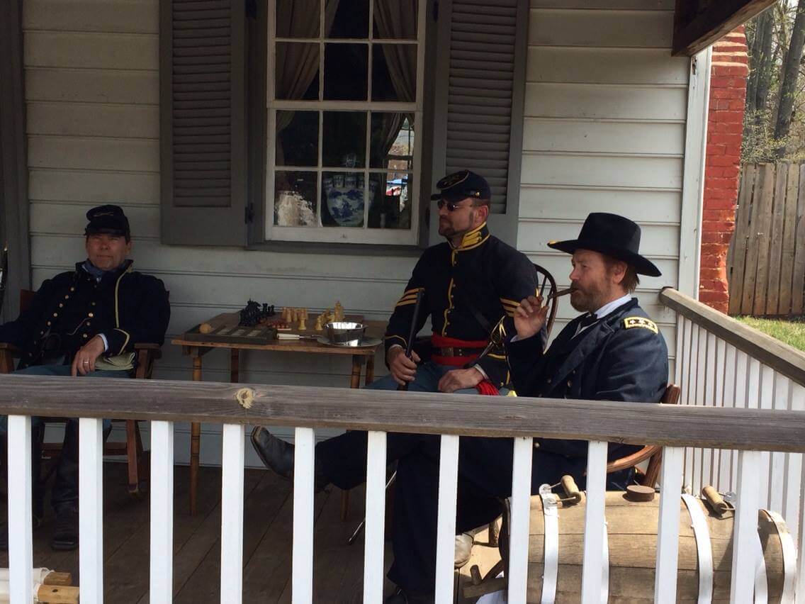 Three men dressed in Civil War-era military uniforms sit on a porch. One on the left is seated casually, another in the middle appears to be lost in thought, and the third on the right is smoking a pipe. A chessboard and other items are on the table behind them.