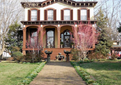 A two-story house with a symmetrical facade, featuring a covered front porch, brown shutters, and trimmed bushes. A pathway lined with budding plants leads to the entrance, flanked by two decorative urns. Trees with pink blossoms are in the front yard.