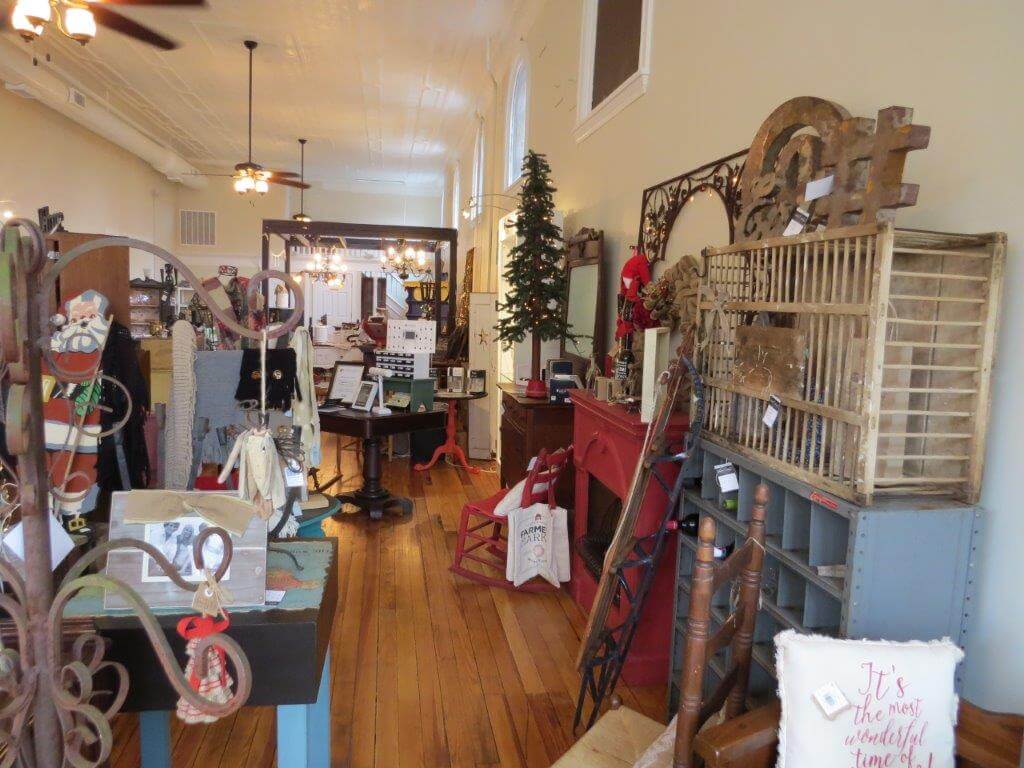 A cozy store is filled with various home decor items and gifts. Vintage furniture, decorative ornaments, and holiday-themed accessories adorn the space. A wooden floor and high ceilings with ceiling fans add to the rustic, inviting ambiance.