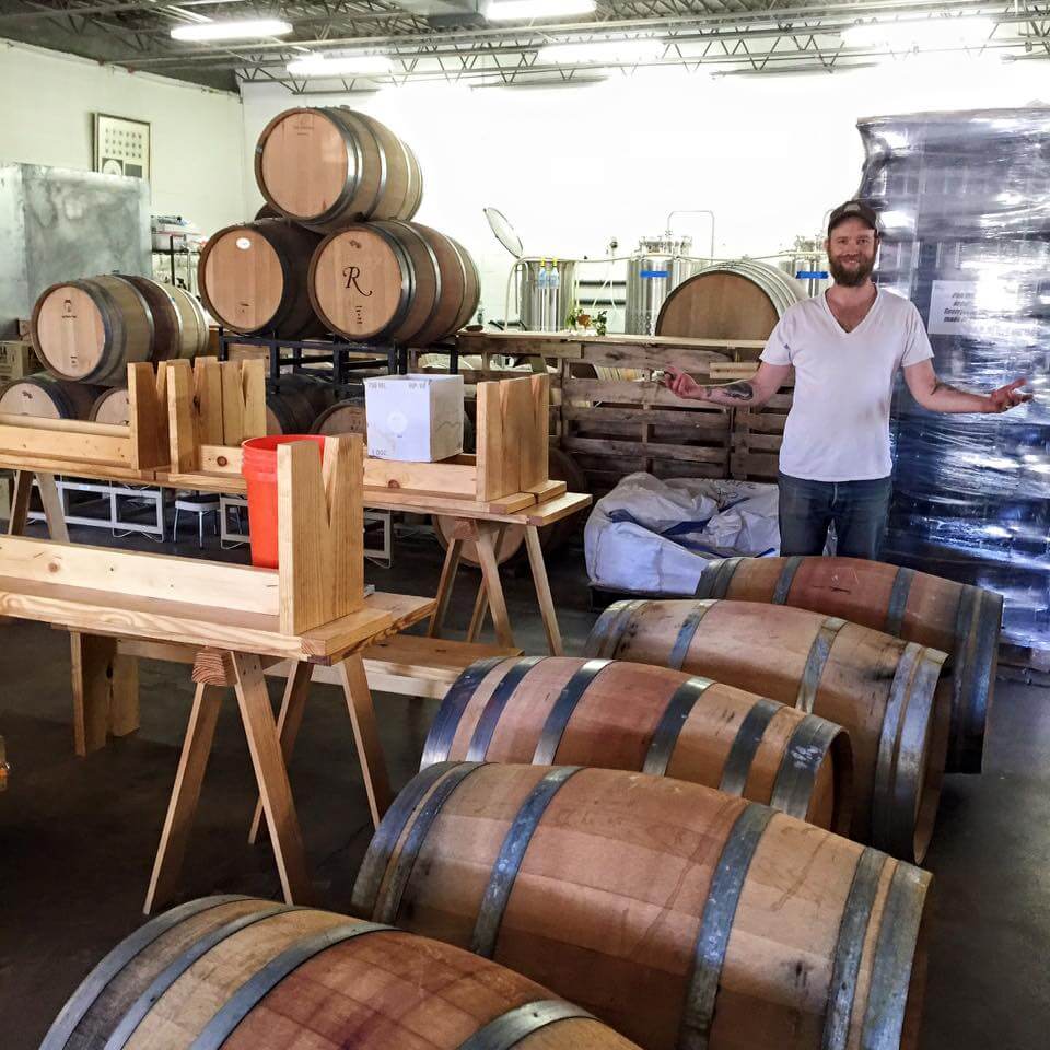 A person with a beard and casual attire stands with outstretched arms in a room filled with wooden barrels, wooden racks, and various industrial equipment. The space appears to be a distillery or winery with supplies and raw materials neatly organized.