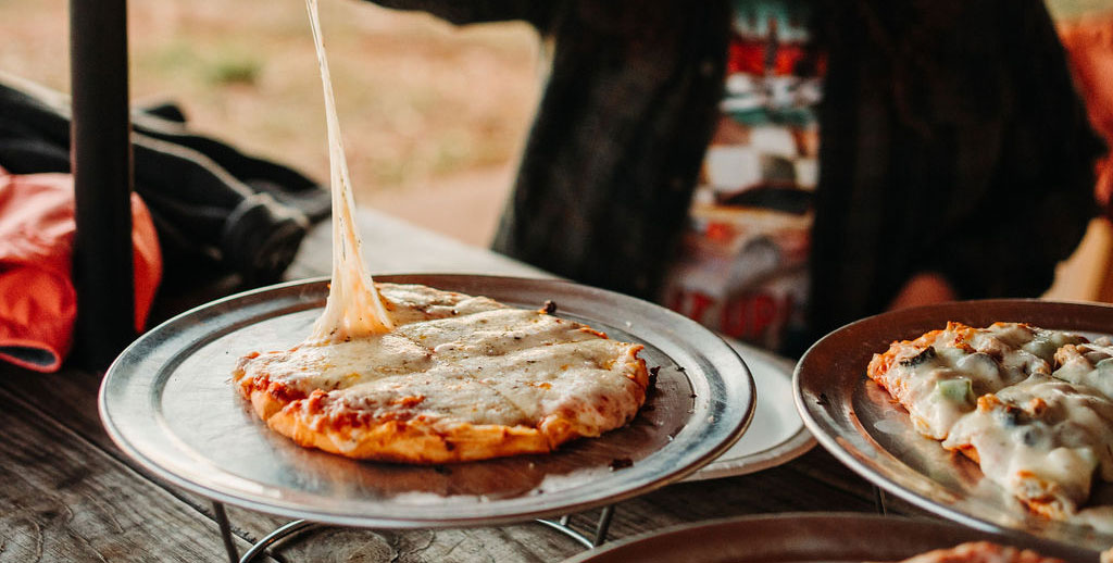 A person lifts a slice of cheese pizza from a metal tray, showcasing a long, stretchy cheese pull. The tray sits on a textured wooden table next to another pizza topped with cheese and vegetables. The background is slightly blurred, focusing on the pizzas.