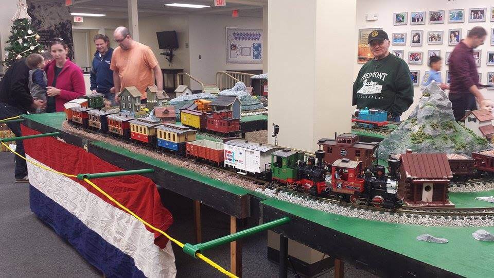 A model train exhibit is displayed on a table adorned with red, white, and blue bunting. Various model trains and buildings are set up on tracks. Several people, including a man wearing a green hat and sweatshirt with text, view the display. A Christmas tree and framed pictures are in the background.