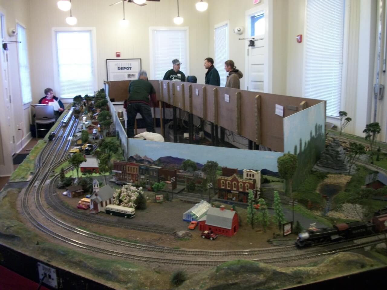 A model railroad display featuring detailed miniature buildings, tracks, and terrain is set up indoors. Several people are gathered around the display, with one person seated at a table in the background. Painted backdrop scenery enhances the model.