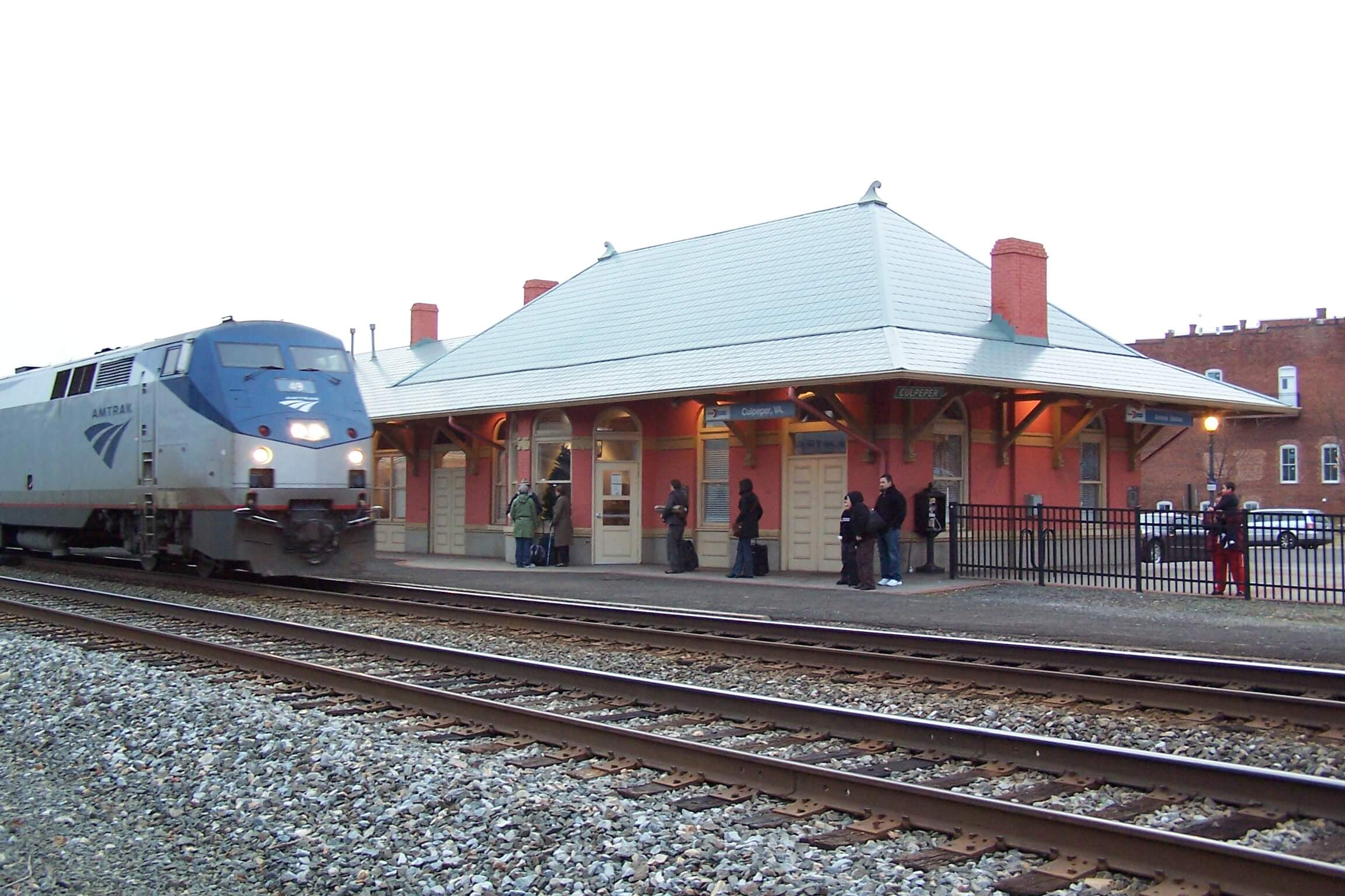 An Amtrak train is approaching a small, quaint railway station with people standing on the platform. The station building has a sloping green roof and a tan exterior with red brick chimneys. There are tracks in the foreground, and brick buildings in the background.