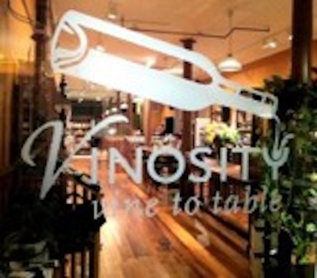 A frosted glass window features a white logo with a wine bottle above the word "VINOSITY" and the tagline "vine to table" below it. Through the glass, a warmly lit, inviting restaurant interior with wooden floors and tables can be seen.