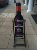 A sidewalk sign resembling a bottle of wine stands on a concrete walkway outside a building. The sign advertises "Adult Beverages & Tasty Treats" at Vinosity and includes a phone number. The wine bottle is deep red with orange and purple accents.