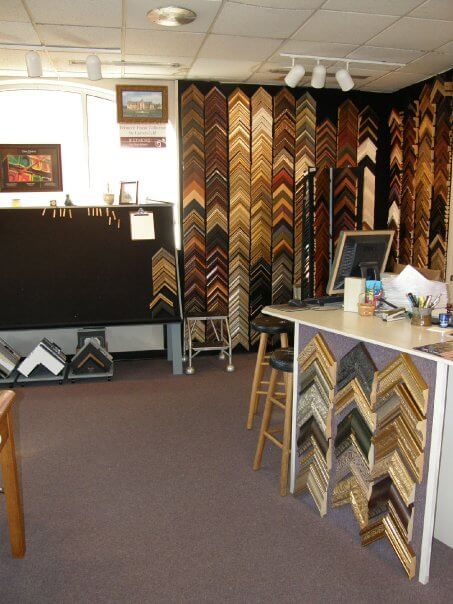 An art framing shop with various picture frame samples displayed on the walls. A counter with design tools and frame corners stands in the foreground. Shelves and racks with frame samples fill the background. The room is well-lit with overhead lighting.