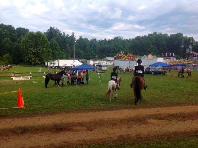 A grassy field with riders on horseback facing away, wearing black jackets with numbers, heading towards canopies and fair rides in the distance. Several horses are tied near a blue canopy, while others roam in the tree-lined background under a cloudy sky.