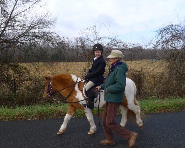 A child wearing a riding helmet and jacket is riding a small brown and white pony on a rural road. An adult in a green jacket and hat walks beside them, holding the pony's reins. Bare trees and fields are visible in the background.