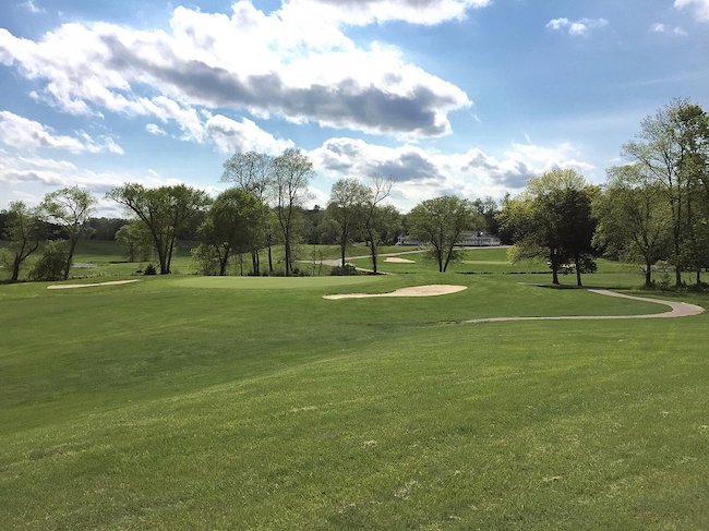 A scenic view of a golf course on a sunny day with various sand traps and a winding cart path. The surrounding landscape features lush green grass and trees scattered across the gently rolling hills under a partly cloudy sky.