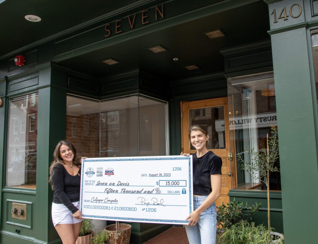Two women stand outside a green storefront named 'Seven,' smiling and holding a large ceremonial check for $15,000. One woman wears a black top and shorts, and the other wears a black top and jeans. The check is dated August 18, 2023, and signed by Calhoun Companies.