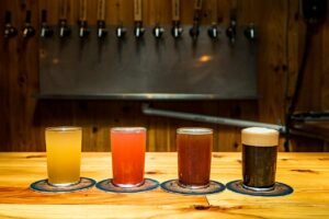 Four glasses of beer, each a different color and type, are placed on individual coasters on a wooden bar. Behind them, a row of beer taps is mounted on the rustic wooden wall. The warmly lit setting is perfect for celebrating Virginia Craft Beer Month in this cozy brewery.