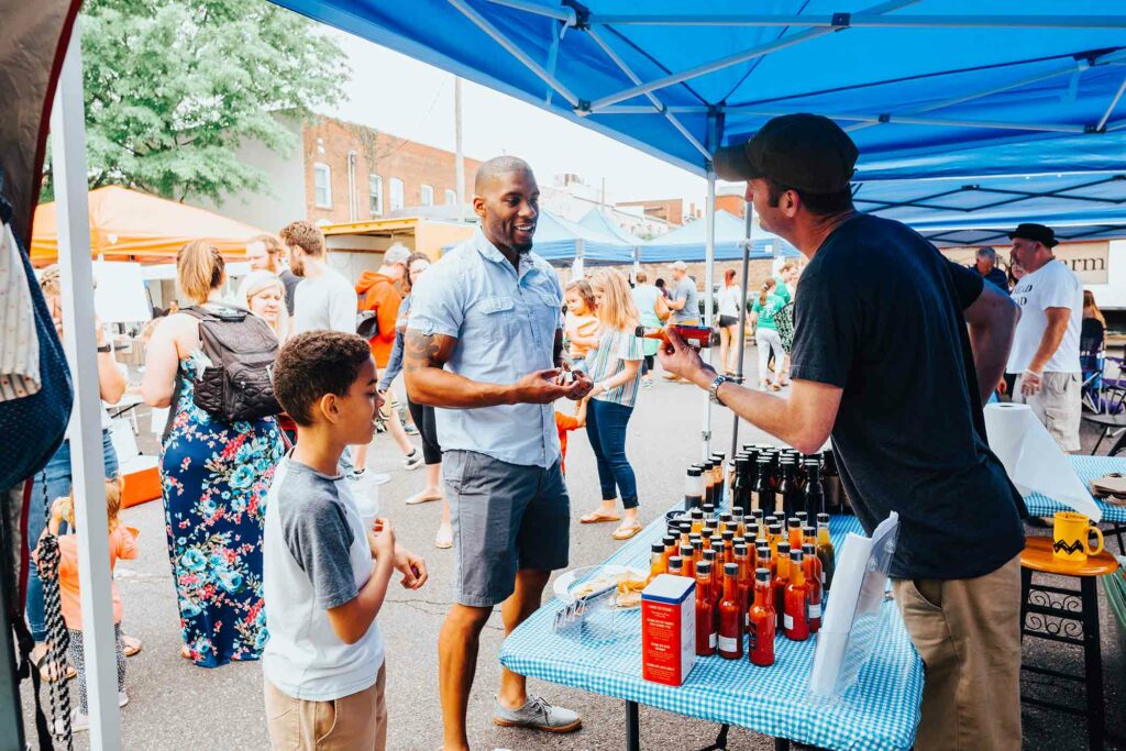 People are gathered at an outdoor market under blue tents, highlighting the summer fun in Culpeper. A vendor is handing a bottle to a man with a child nearby while various bottles are displayed on the table. Shoppers and booths dot the background, creating a friendly and vibrant atmosphere.