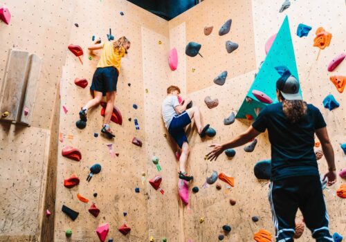 Two climbers, one in a yellow shirt and the other in white, scale a colorful indoor climbing wall. A third person in a black shirt and cap watches them from below, offering guidance. Various holds and grips of different shapes and colors are attached to the wall.