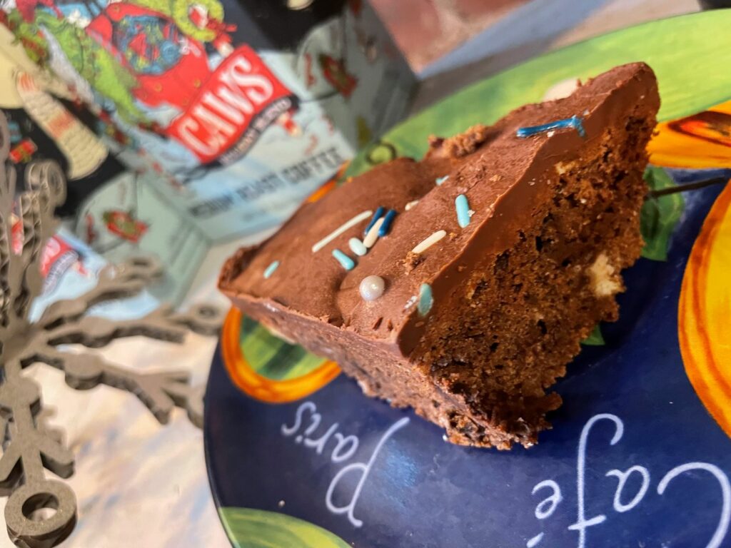 A slice of chocolate cake with chocolate frosting and blue and white sprinkles sits on a colorful plate that reads 'cafe Paris.' In the background, among other sweet treats, there's a festive-themed tin can. A snowflake decoration is partially visible to the left.