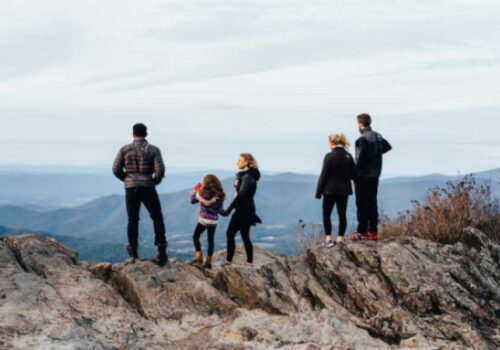 Five people, including three adults and two children, stand on a rocky outcrop overlooking a scenic mountain range. They face away from the camera, taking in the expansive view under a cloudy sky. Some wear jackets and hiking shoes, indicating a outdoor adventure.