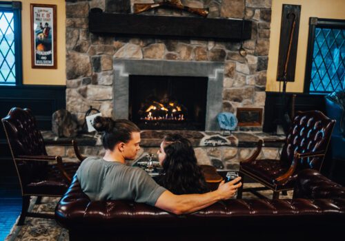 A couple with drinks sits on a brown leather couch, facing a stone fireplace with a warm fire. The rustic room has dark wooden panels, framed windows, and a poster on the wall. They appear cozy and comfortable in the homey setting.