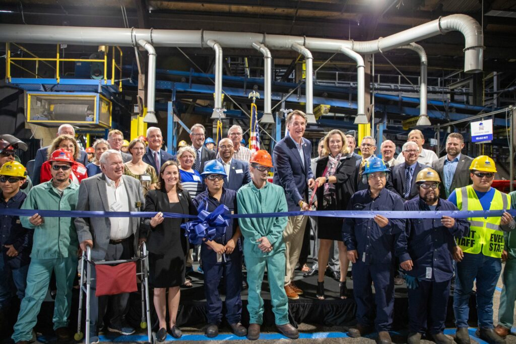 A diverse group of people, including construction workers in safety gear and several individuals in business attire, are gathered in a factory setting for a ribbon-cutting ceremony. The group is smiling and posing, with a large blue ribbon being cut at the center.