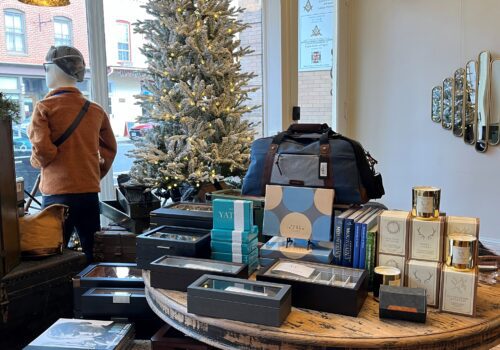 A cozy store with warm wooden floors, featuring a decorated Christmas tree near the window. A table displays various gift boxes, skincare products, and a stylish bag perfect for holiday shopping. A person in a brown jacket and beanie stands by the window, looking outside.