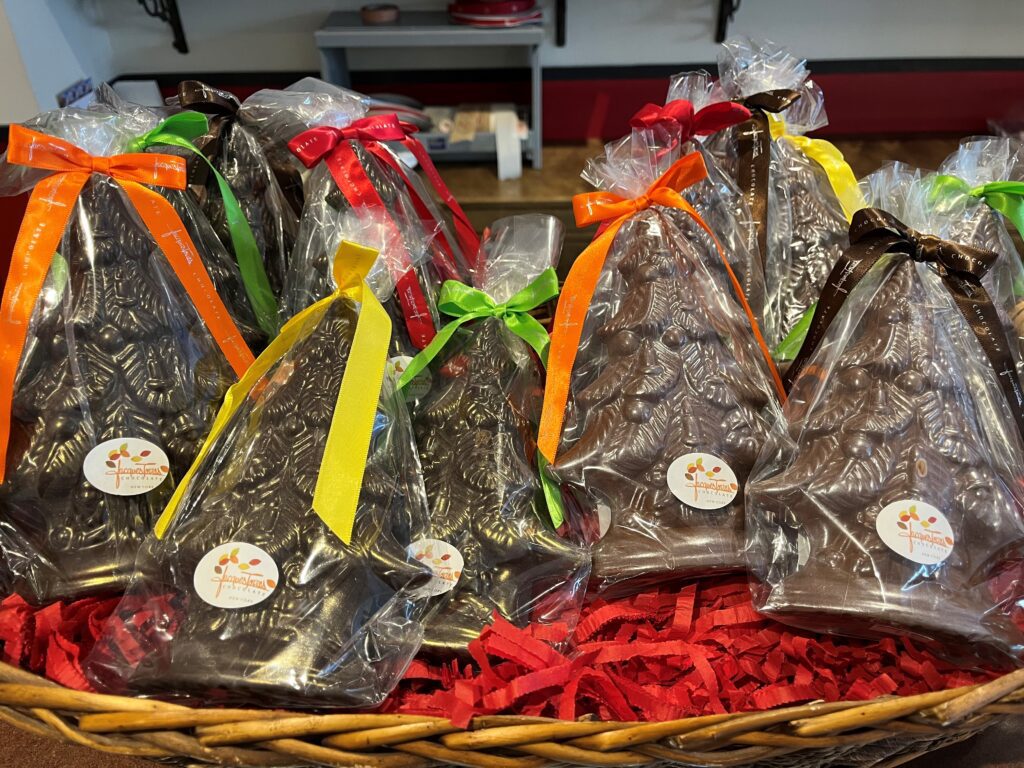 A basket filled with chocolate Christmas trees, wrapped in clear plastic and tied with colorful ribbons. Each tree has a small white sticker with an orange logo and text on it. Shredded red paper lines the basket, adding to the festive feel of holiday shopping. A shop counter is in the background.