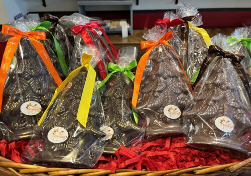 A basket filled with chocolate Christmas trees, wrapped in clear plastic and tied with colorful ribbons. Each tree has a small white sticker with an orange logo and text on it. Shredded red paper lines the basket, adding to the festive feel of holiday shopping. A shop counter is in the background.