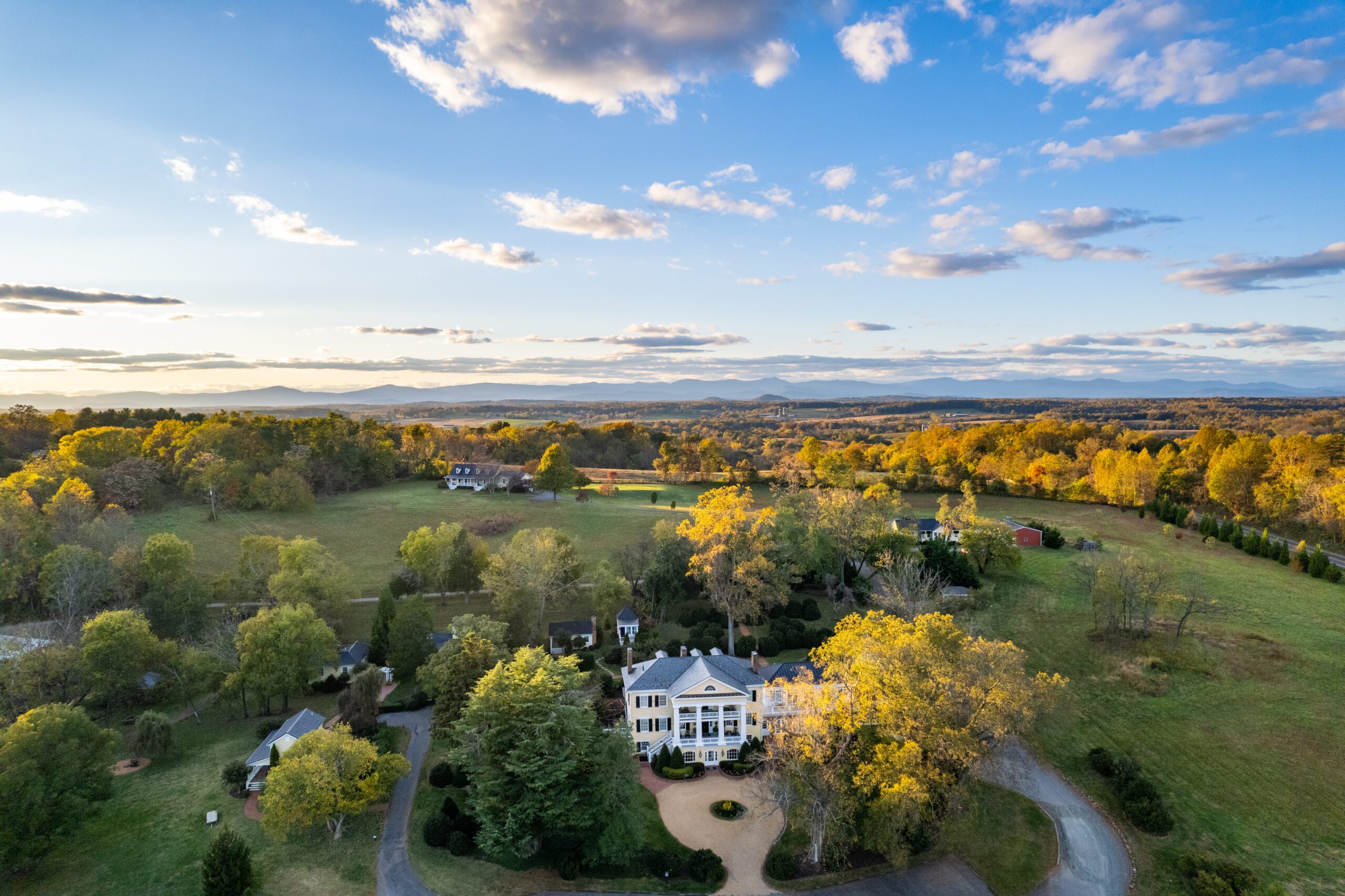 Aerial view of a large white house with a circular driveway and surrounding green lawns and trees under a blue sky with scattered clouds. Rolling hills and distant mountains are visible in the background.