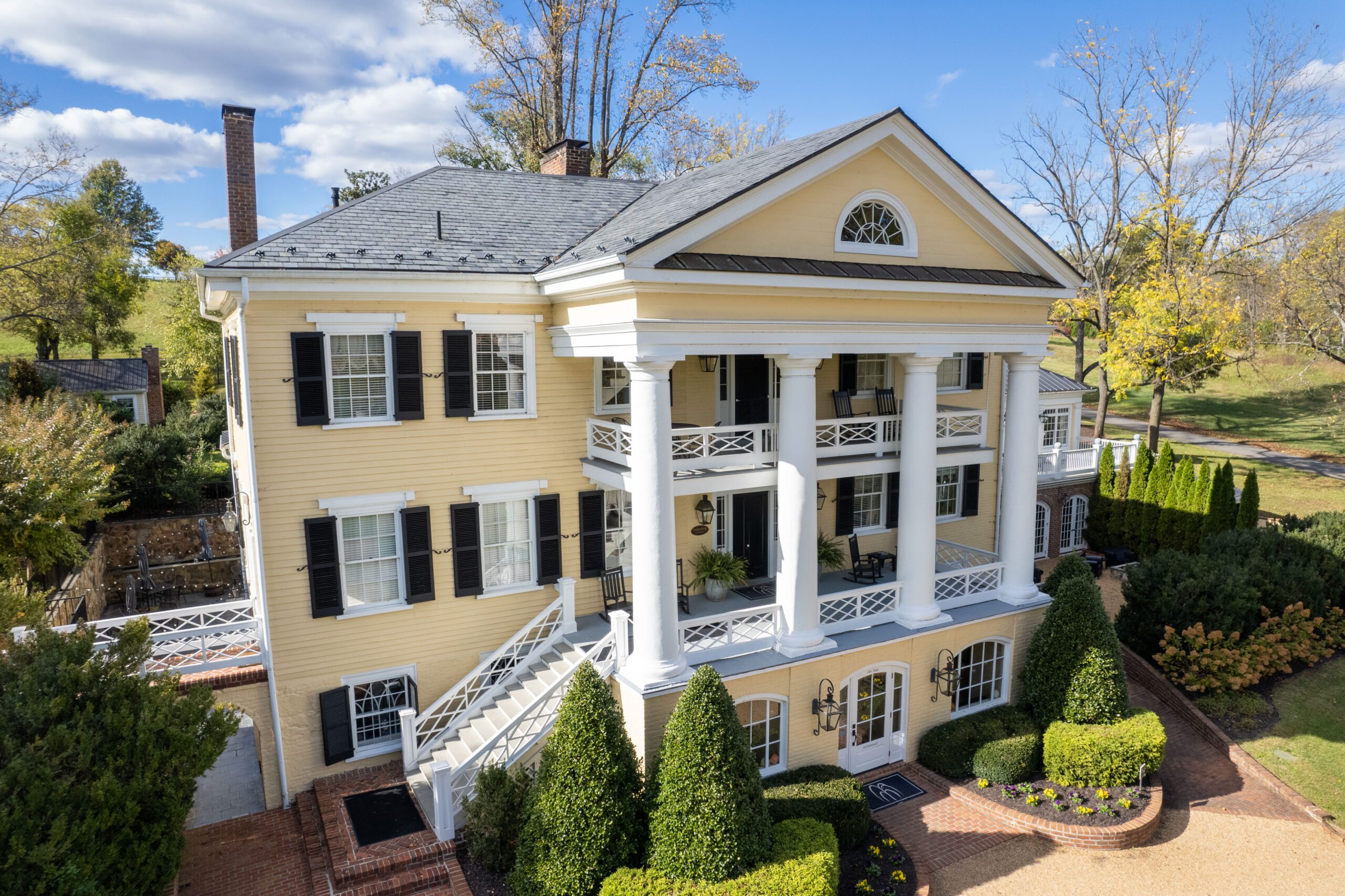 A large, elegant two-story house painted in a soft yellow stands against a backdrop of tall trees and a sunny sky. It features white columns, black shutters, and a grand front entrance with a staircase on each side. Manicured shrubs and hedges line the walkway to the entrance.