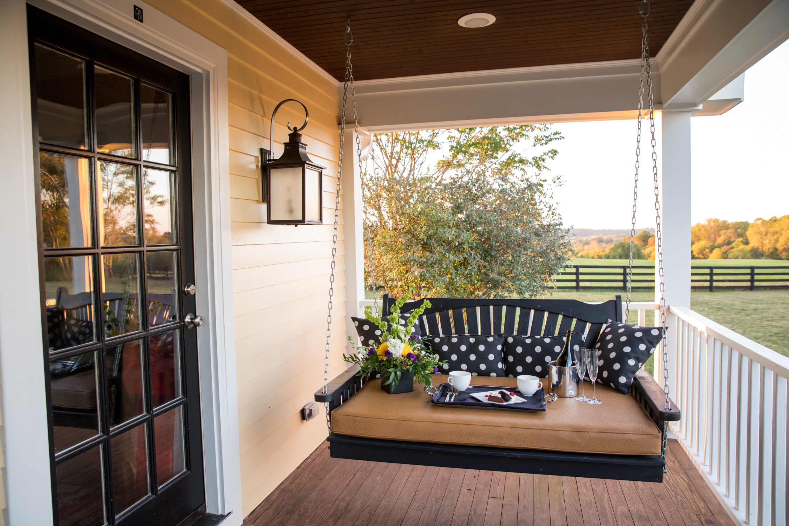 A cozy porch features a black wooden swing with beige cushions and black polka dot pillows. A tray with a teapot, cups, and a vase of flowers sits on the swing. The porch has a black-framed glass door, a wall-mounted lantern, and a view of trees and fields.