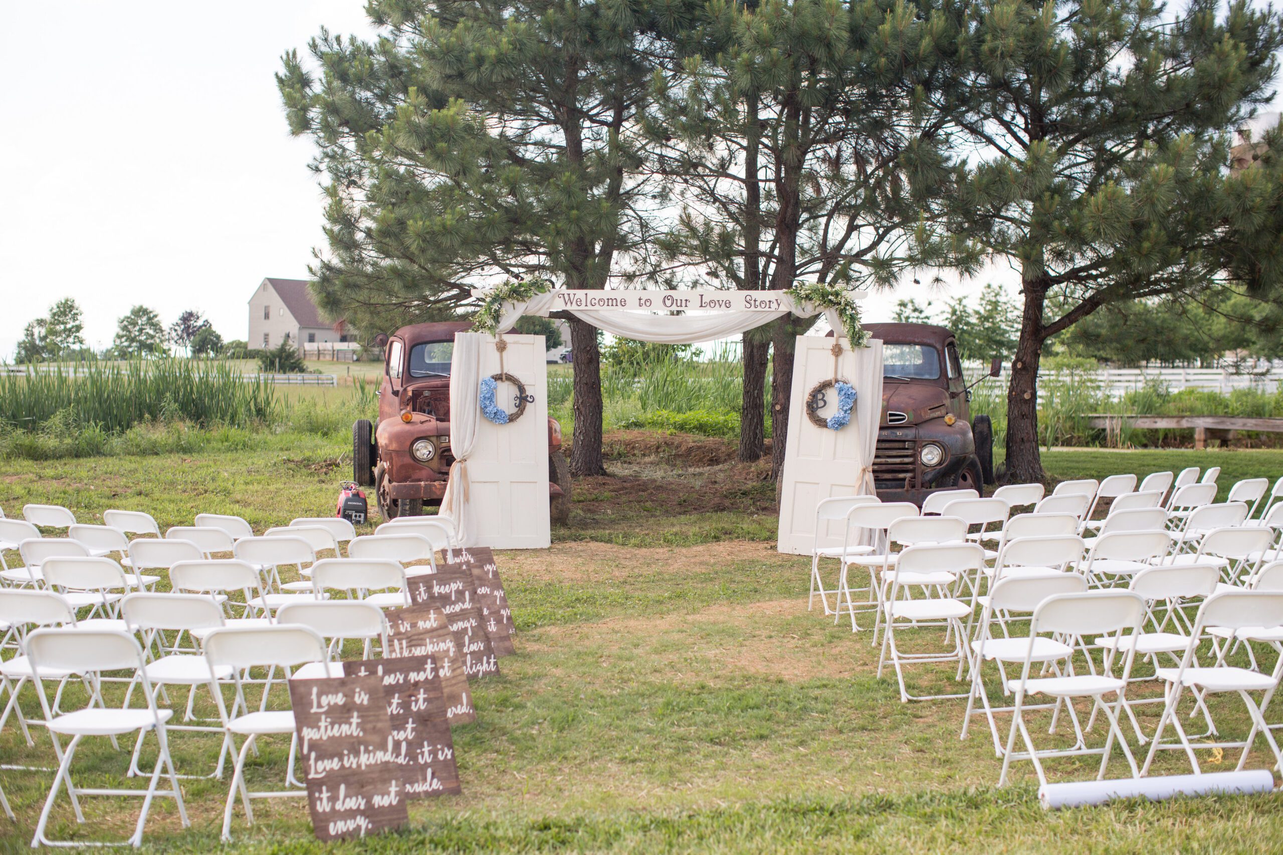 An outdoor wedding setup featuring rows of white chairs facing an aisle flanked by antique doors adorned with wreaths. A banner reading 